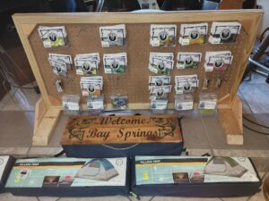 bt 4 Display board with assorted fishing lines, a wooden "Welcome Bay Springs" sign, and three boxed tents on the floor.