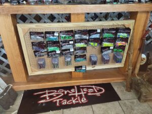 bt 9 Fishing lures and tackle displayed on a pegboard stand above a BoneHead Tackle floor mat in a store.