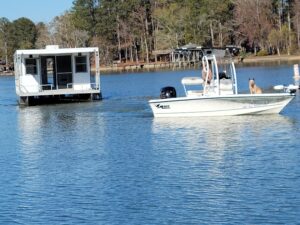 towing 1 A small white motorboat with two dogs and a person passes a houseboat on a calm lake, surrounded by trees.