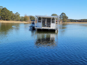 towing 2 A small houseboat is being towed on a calm lake surrounded by trees under a clear blue sky.