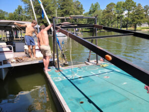 towing 4 Two men work shirtless on a docked boat with ropes and metal beams on a sunny day by the lake.